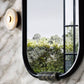 Round mirror with black frame on a marble wall, reflecting greenery outside.
