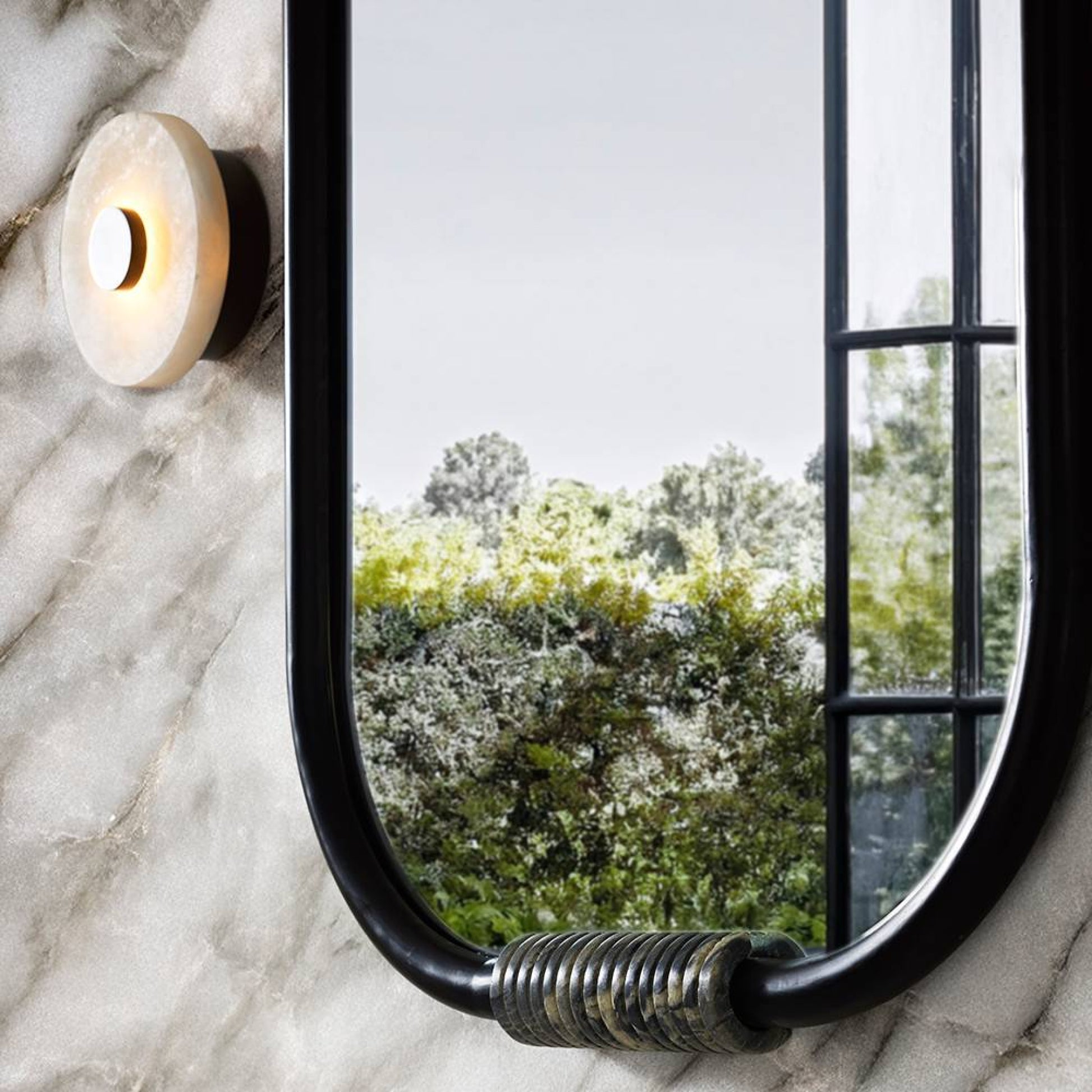 Round mirror with black frame on a marble wall, reflecting greenery outside.