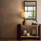 Decorative console table with lamp, mirror, and sculptures against a beige wall.