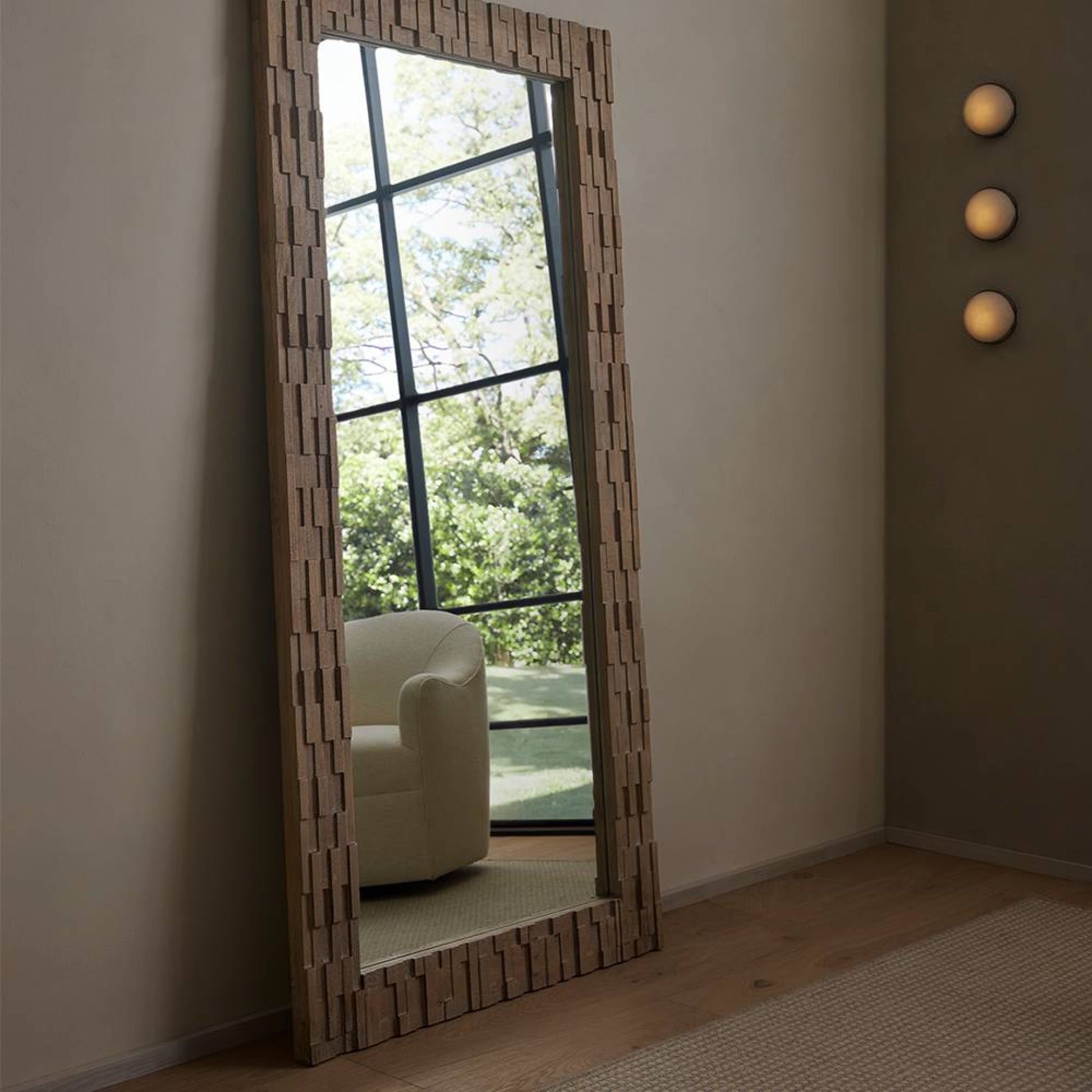 Large decorative mirror with geometric frame in a room with a white armchair and greenery outside.