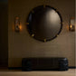 Round mirror with decorative frame on a textured wall with wall sconces.