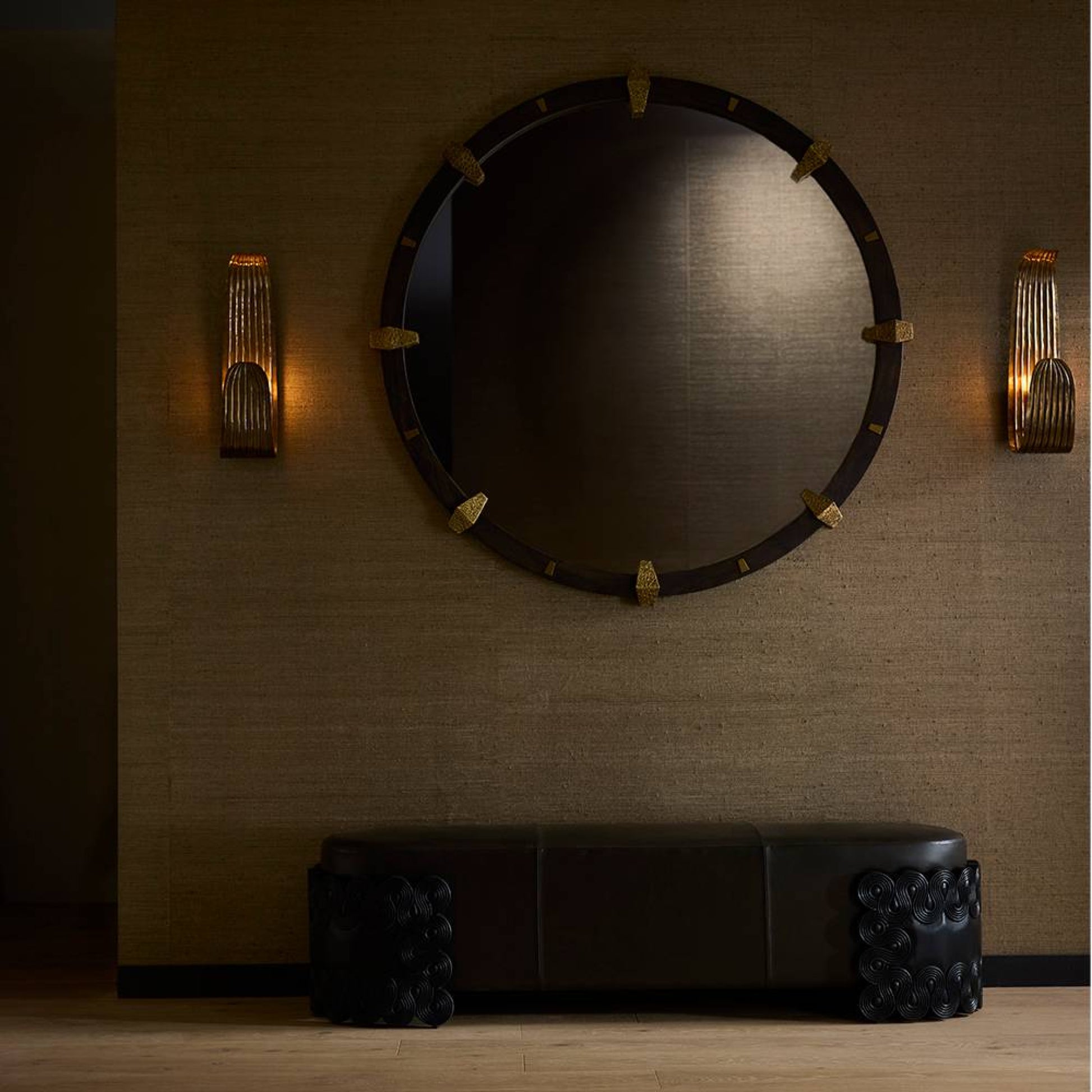 Round mirror with decorative frame on a textured wall with wall sconces.