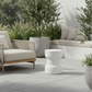 A modern outdoor lounge chair with woven beige wicker-style arms and seat cushion, paired with a small wooden side table and a white sculptural side stool on a light stone patio. The scene includes potted olive trees, a built-in bench in the background, and soft natural lighting.