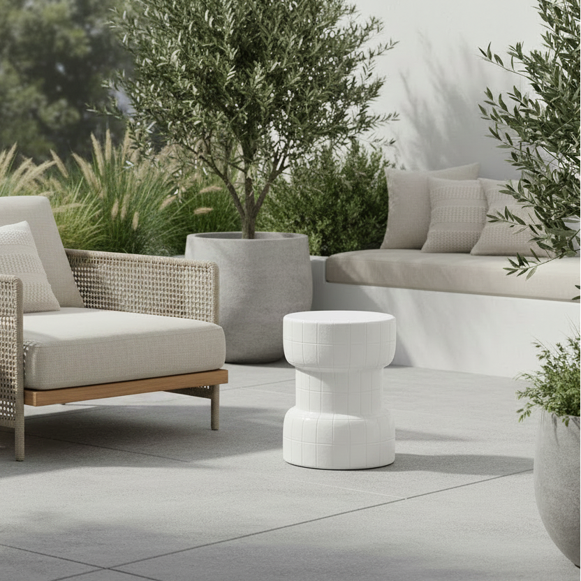 A modern outdoor lounge chair with woven beige wicker-style arms and seat cushion, paired with a small wooden side table and a white sculptural side stool on a light stone patio. The scene includes potted olive trees, a built-in bench in the background, and soft natural lighting.