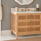 A bathroom vanity cabinet with three drawers featuring woven cane panels, a white marble countertop, a white sink, and a brass faucet. 
