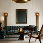 A styled interior scene featuring a teal tufted sofa, a round dark wood coffee table with gold accents, a woven wood armchair, a wall mirror, and a large brass pendant light against a warm off-white wall.