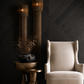 A cream-colored wingback armchair in a styled interior setting with a dark wood chevron wall, two tall woven pendant lights, and a bronze side table holding a black ceramic vase with dried branches.