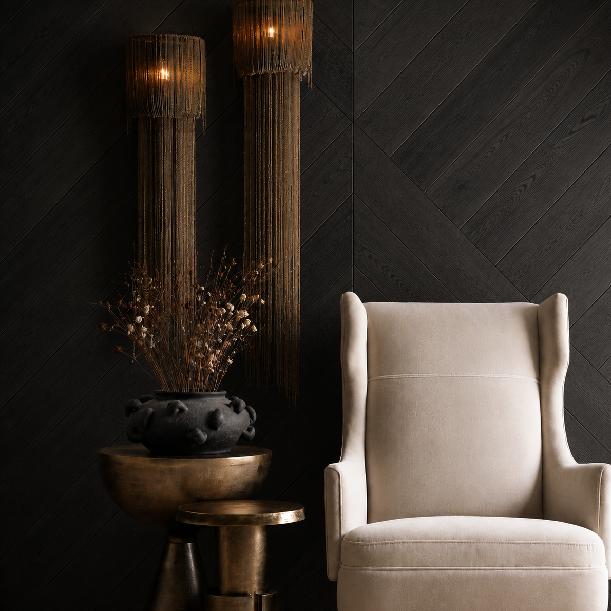 A cream-colored wingback armchair in a styled interior setting with a dark wood chevron wall, two tall woven pendant lights, and a bronze side table holding a black ceramic vase with dried branches.