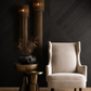 A cream-colored wingback armchair in a styled interior setting with a dark wood chevron wall, two tall woven pendant lights, and a bronze side table holding a black ceramic vase with dried branches.