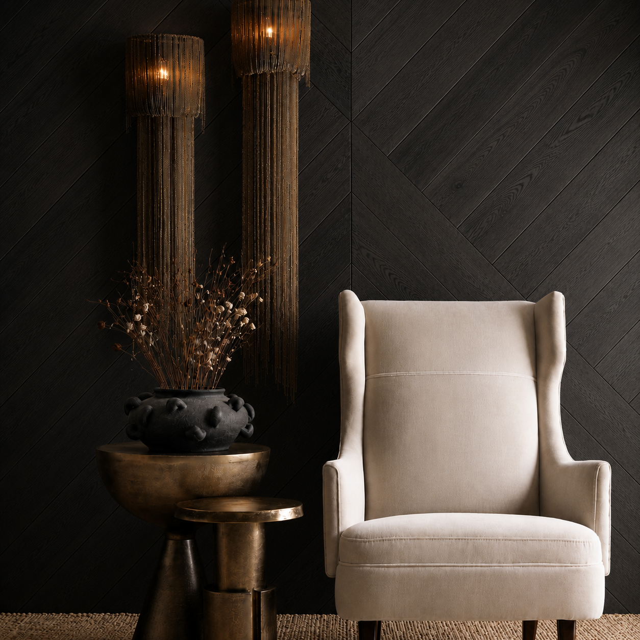 A cream-colored wingback armchair in a styled interior setting with a dark wood chevron wall, two tall woven pendant lights, and a bronze side table holding a black ceramic vase with dried branches.