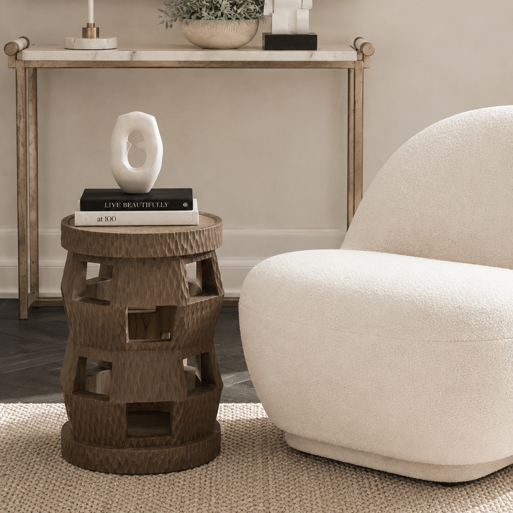 A styled interior scene showing a round woven rattan side table with geometric cutout openings, styled with books and a white sculptural vase. Next to it is a modern curved white armchair, with a console table, lamp, and framed art in the background.