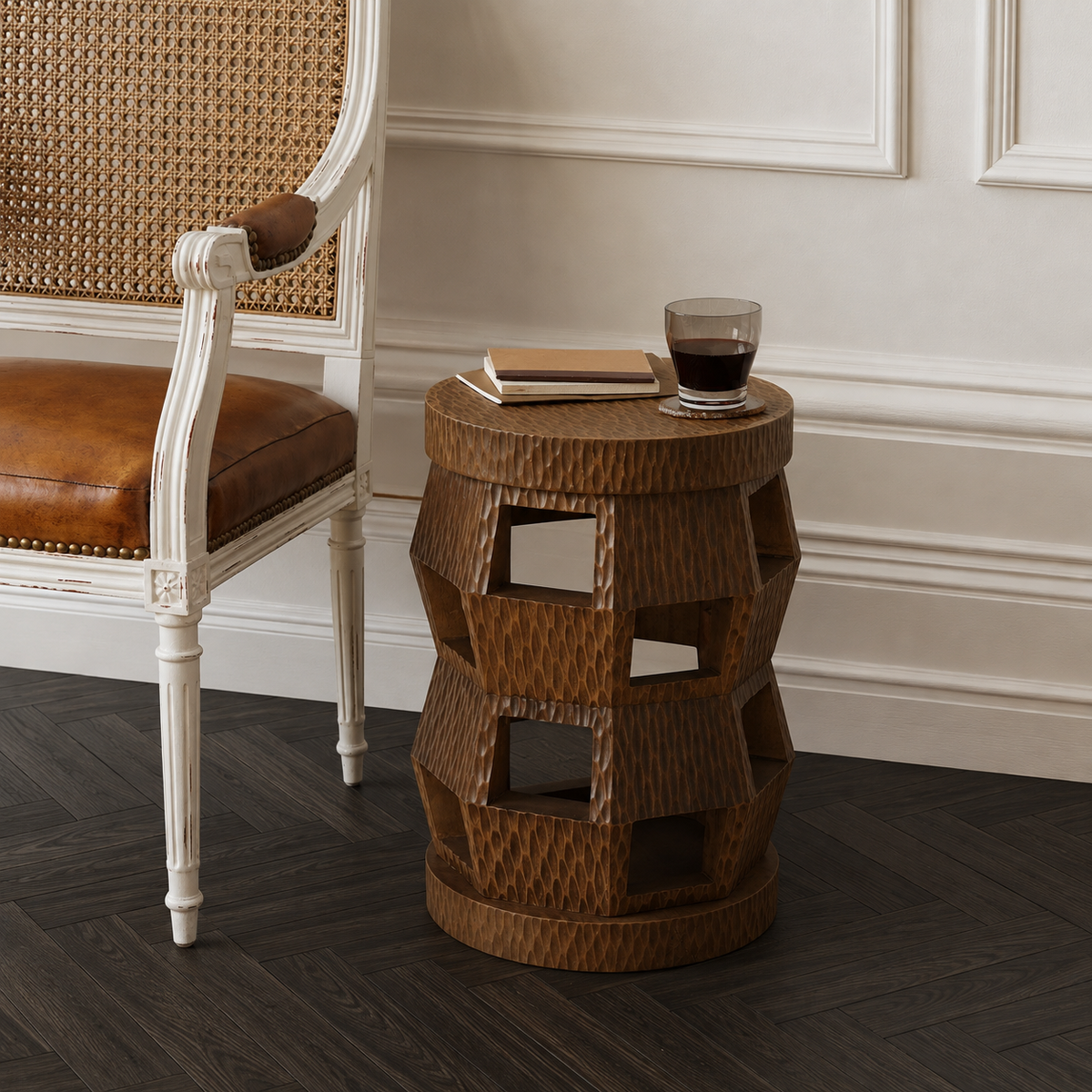 A round, geometric side table made of dark brown woven rattan with open cuboid cutouts, styled in a room next to a white armchair with a brown leather seat. A glass of wine, a notebook, and a book rest on top of the table.