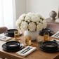 A styled dining table setting with black ceramic plates, matching black bowls, amber-tinted glass cups, white napkins, and silver cutlery, centered around a large white vase filled with white roses and cotton stems.