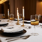 A styled dining table setting with white tablecloths, black plates, folded white napkins, silver cutlery, amber-colored wine glasses, and multiple lit white taper candles in gold and bronze holders in a warm indoor environment.